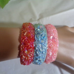 May include: Three iridescent plastic bangles with a floral design. The bangles are pink, blue, and red.