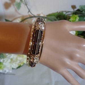 May include: Gold bangles with detailed designs. One bangle has a black central section with vertical gold lines and sparkling embellishments. The other is decorated with small, clear gemstones. A classic piece of jewellery.
