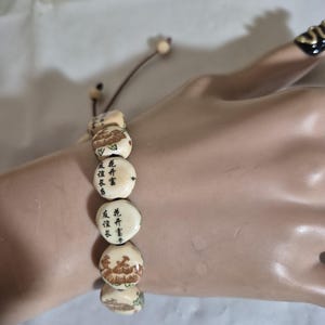 May include: A bracelet featuring off-white, rounded beads with brown floral illustrations and black Chinese characters. The beads are strung on a thin brown cord with adjustable knots and small, round, light-colored wooden accent beads at the ends.