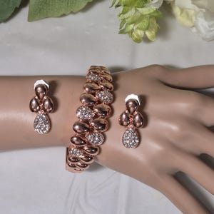 May include: Rose gold-toned jewelry set featuring a bracelet and matching earrings. The bracelet has a series of oval links, some encrusted with clear crystals. The earrings have a teardrop shape with crystal accents, creating a coordinated look.