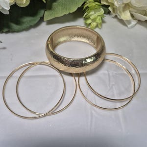 May include: A gold-toned jewelry set featuring a wide, textured bangle bracelet and three thin, smooth bangles. The bracelets are arranged on a white surface, with floral accents in the background.