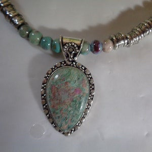 May include: A silver necklace with a green and pink stone pendant. The necklace is made of silver beads and has a silver clasp.