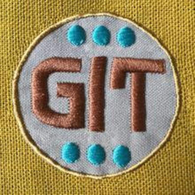 Op de afbeelding: Een blauwe en bruine geborduurde patch met de letters "GIT" in bruin en vier turquoise stippen rond de rand.