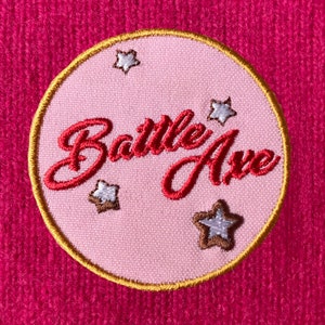 May include: A pink fabric patch with a gold border and the text "Battle Axe" in red embroidery. The patch is decorated with five white stars with brown outlines.