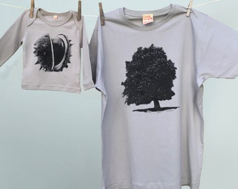 Può includere: Due camicie grigio chiaro appese a un filo. Una è una tutina a maniche lunghe per neonati con una grafica in bianco e nero di una castagna. L'altra è una t-shirt con una grafica di un albero nero.