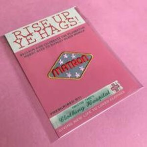 Op de afbeelding: Een roze en witte kaart met de tekst "Rise Up Ye Hags!" en "Prescribed Clothing Hospital" in rood en zwart. Een blauw en wit ruitvormig patch met de tekst "Matron" in rood is aan de kaart bevestigd.