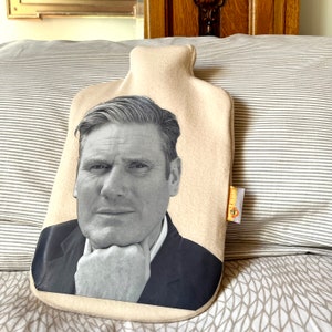 May include: A beige fabric hot water bottle with a black and white photo of a man's face on the front. The man is wearing a suit jacket and has his hand on his chin.