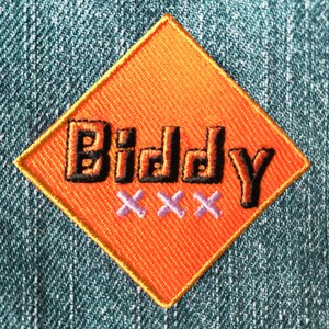 Op de afbeelding: Oranje ruitvormige patch met het woord "Biddy" in zwarte omrande letters en drie paarse "X" eronder.