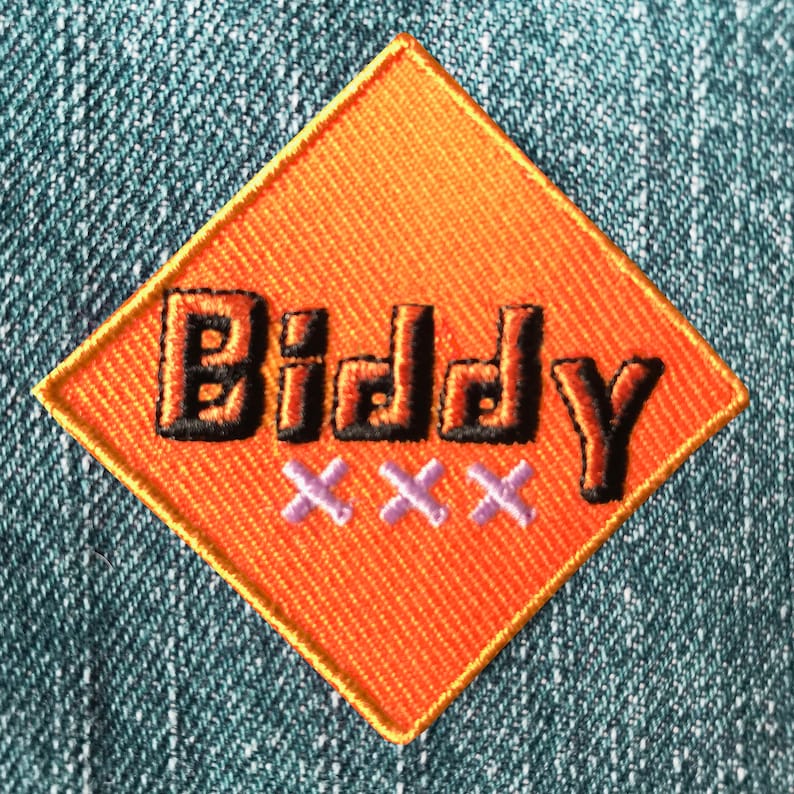 Op de afbeelding: Oranje ruitvormige patch met het woord "Biddy" in zwarte omrande letters en drie paarse "X" eronder.