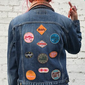 Op de afbeelding: Een blauwe denimjas met verschillende geborduurde patches. De patches hebben kleurrijke ontwerpen en tekst, waaronder "Battle Ave", "Biddy", "Matron", "Misery Guts", "codger", "Old Fogy", "Hag", "Fish Wife", "Crone" en "Git".