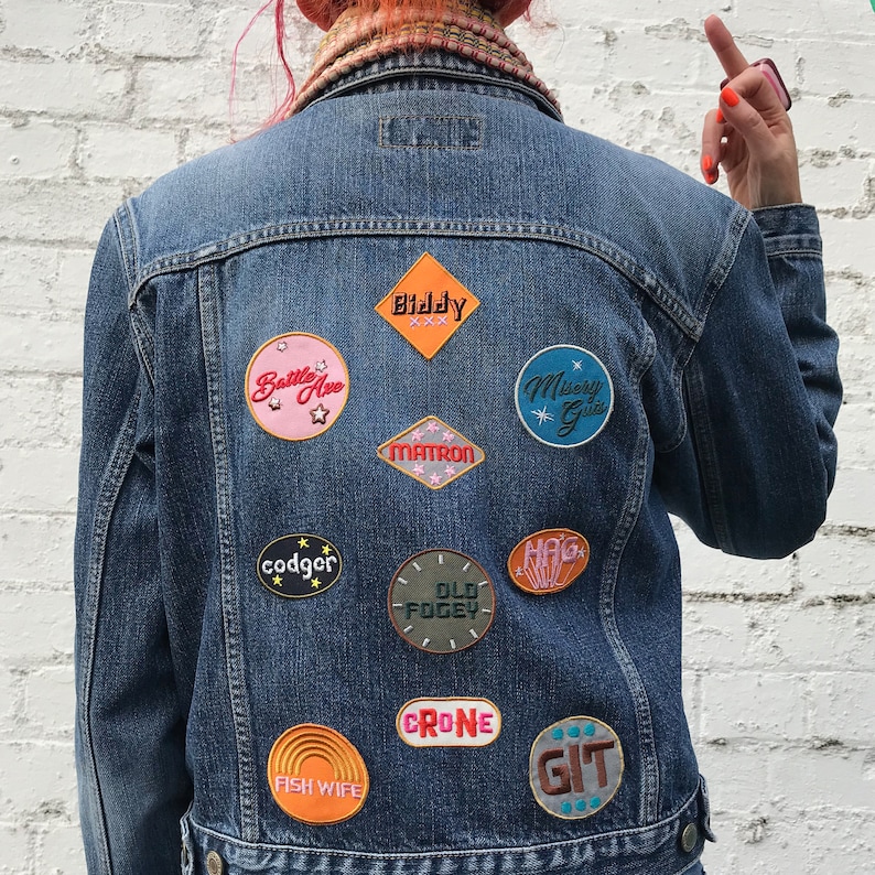 Op de afbeelding: Een blauwe denimjas met verschillende geborduurde patches. De patches hebben kleurrijke ontwerpen en tekst, waaronder "Battle Ave", "Biddy", "Matron", "Misery Guts", "codger", "Old Fogy", "Hag", "Fish Wife", "Crone" en "Git".