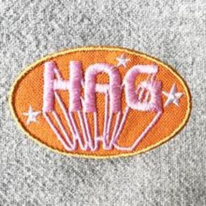 May include: Embroidered patch with the word "HAG" in pink, outlined in white, on an orange oval. White star accents are on either side of the text. The patch has a yellow border and is on a textured gray background.