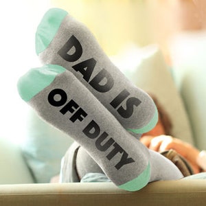 Op de afbeelding: Grijze sokken met een mintgroene hiel en teen. De sokken hebben de tekst "Dad is Off Duty" in zwart erop gedrukt.