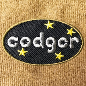 Op de afbeelding: Een geborduurde patch met het woord "codger" in het wit, gestikt op een zwarte ovaal. De patch is omgeven door een gouden rand en heeft vier gele steraccenten. De achtergrond is een getextureerde beige stof.