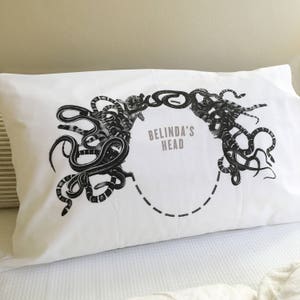 Könnte beinhalten: Weißer Kissenbezug mit einer schwarz-weißen Illustration von Schlangen, die einen Kreis um den Text "BELINDA'S HEAD" bilden.