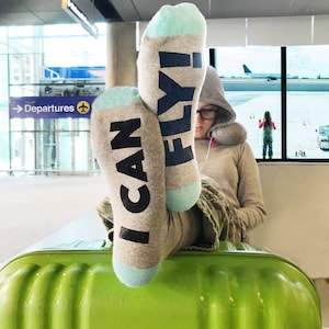 Può includere: Un paio di calzini grigi con dettagli blu. Un calzino dice "I CAN" e l'altro dice "FLY!" I calzini sono indossati da una persona seduta su una valigia verde in un aeroporto.
