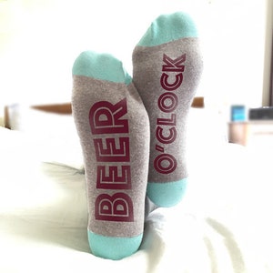 May include: A pair of gray socks with blue heels and toes. One sock says "Beer O'Clock" and the other says "Beer".
