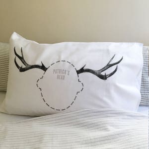 Könnte beinhalten: Weißes Kopfkissen mit einem schwarzen Geweih-Design und dem Text "PATRICK'S HEAD" in Grau. Das Kissen ist rechteckig und liegt auf einem Bett mit einer gestreiften Bettdecke. Das Geweih ist symmetrisch und detailliert.