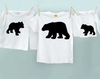 May include: Three white t-shirts with black bear silhouettes hanging on a clothesline. The shirts vary in size, with one long-sleeved and two short-sleeved, each featuring a different bear silhouette. The background is a light blue.