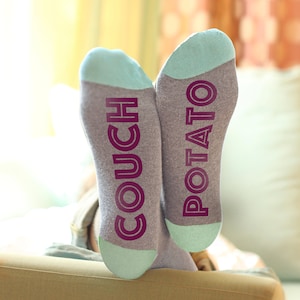 Puede incluir: Un par de calcetines grises con talón y puntera azul claro. Los calcetines tienen las palabras "COUCH POTATO" escritas en ellos en una fuente morada.