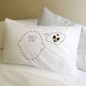 Könnte beinhalten: Weißes Kissen mit einer grauen Umrandung eines Kopfes und dem Text "JAKE'S HEAD". Eine weiße Wolke mit schwarzem Umriss enthält einen schwarz-weißen Fußball.