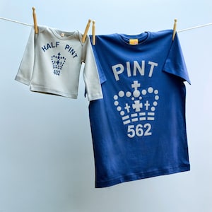 以下が含まれることがあります： 洗濯物干しにかけられた2枚のTシャツ。小さい方のTシャツは薄いグレーで、「HALF PINT 402」と王冠のグラフィックが書かれている。大きい方のTシャツは青で、「PINT 562」と王冠のグラフィックが書かれている。