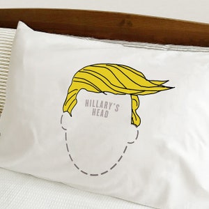 Könnte beinhalten: Weißes Kopfkissen mit gelber Cartoon-Frisur und dem Text "HILLARY'S HEAD" in Grau. Das Kissen hat ein minimalistisches Design, geeignet für ein Bett oder Sofa.