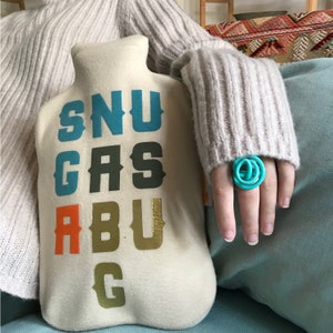 May include: A white fabric hot water bottle with the word "SNUGBUG" printed in colorful letters. The letters are in different colors, including blue, green, orange, and gold.