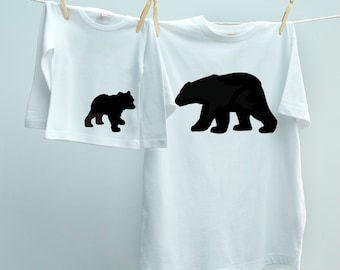 May include: Two white t-shirts hanging on a clothesline. The larger shirt has a black bear silhouette printed on the front. The smaller shirt has a smaller black bear silhouette printed on the front.