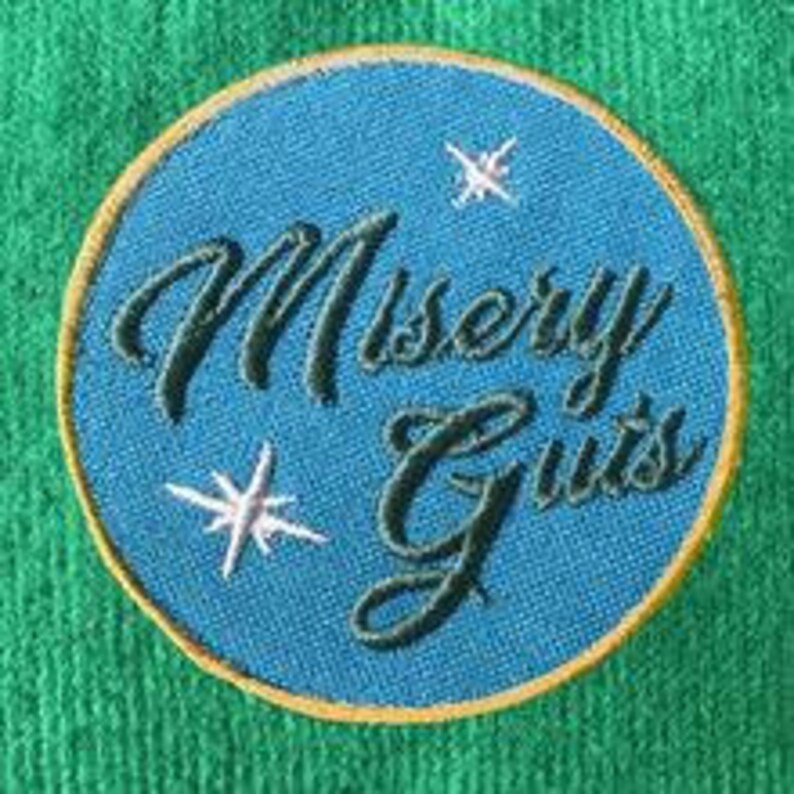 Op de afbeelding: Een blauwe en gouden geborduurde patch met de tekst "Misery Guts" in een cursief schrift. Er zijn twee witte sterren op de patch.