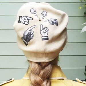 Puede incluir: Un gorro de punto beige con gráficos de manos apuntando en blanco y negro. El gorro lo lleva una persona con el pelo largo y rubio en una trenza.