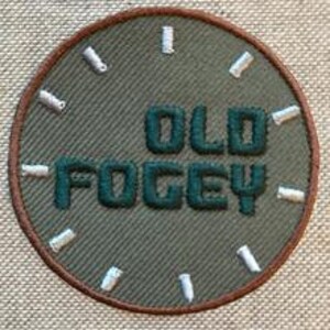 Op de afbeelding: Een groene geborduurde patch met de woorden "OLD FOGY" in witte stiksels op een bruine cirkel.