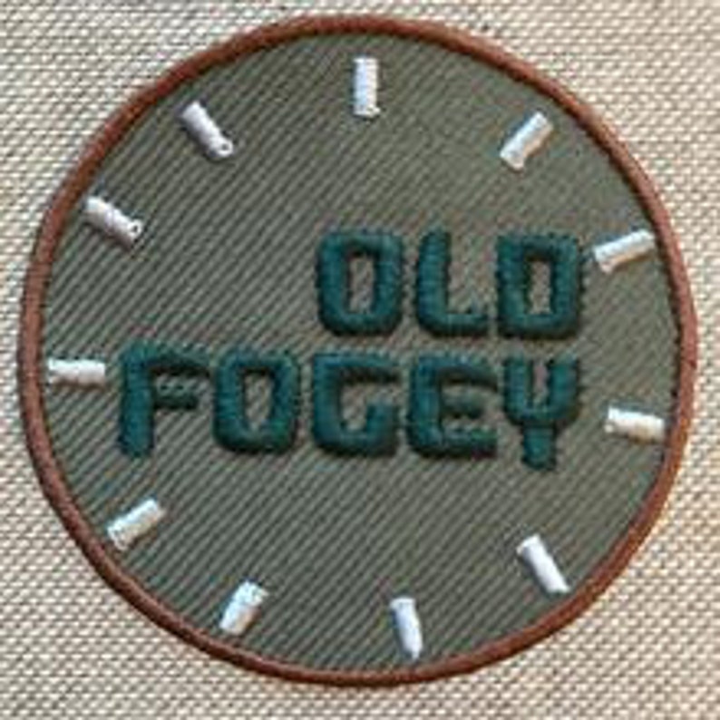 Op de afbeelding: Een groene geborduurde patch met de woorden "OLD FOGY" in witte stiksels op een bruine cirkel.