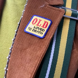 Könnte beinhalten: Eine braune Cordjacke mit einem blau-gelb gestreiften Patch, auf dem "OLD & LOVED TO DEATH" steht.