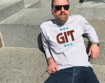 May include: A man wearing a grey long-sleeved t-shirt with the word "GIT" printed in brown letters and surrounded by turquoise dots. He is wearing blue jeans and sunglasses.