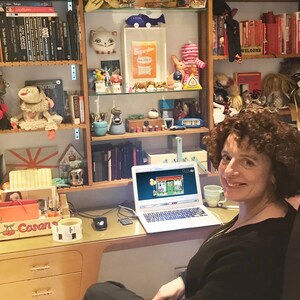 Op de afbeelding: Een vrouw zit aan een bureau met een laptop die open staat met een spel. Het bureau is vol met boeken, speelgoed en andere spullen. De vrouw glimlacht en kijkt naar de camera.