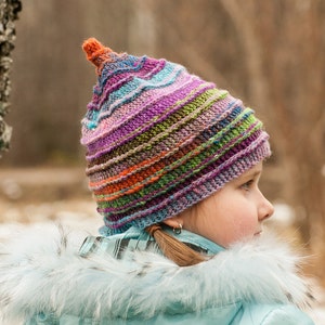 Peut inclure: Une jeune personne portant un bonnet tricoté rayé de couleurs vives avec un sommet pointu. Le bonnet est fait de plusieurs couleurs, dont le violet, le bleu, le vert, l'orange et le brun. La personne porte une veste bleu clair avec un col en fourrure blanche.