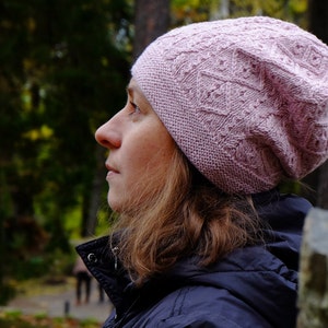 Peut inclure: Une femme portant un bonnet en tricot rose avec un motif en tricot torsadé. Elle se tient dans une forêt et regarde de côté.