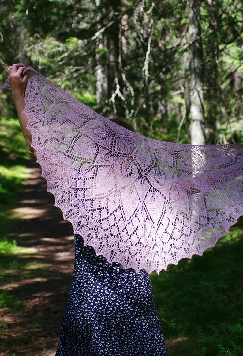 May include: A delicate, lacy shawl in a soft purple hue, draped over a person's shoulders in a forest setting. The shawl features intricate floral patterns and a scalloped edge.