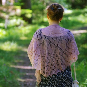 May include: A woman wearing a delicate, lacy, light pink shawl with a floral pattern. The shawl is draped over her shoulders and hangs down to her waist. She is standing in a forest setting with green trees and foliage in the background.