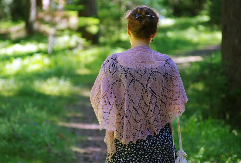 May include: A woman wearing a delicate, lacy, light pink shawl with a floral pattern. The shawl is draped over her shoulders and hangs down to her waist. She is standing in a forest setting with green trees and foliage in the background.