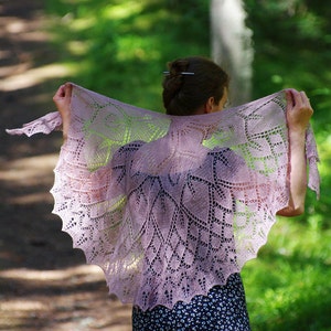 May include: A woman wearing a delicate, lacy, light purple shawl with a floral pattern. The shawl is draped over her shoulders and is flowing in the wind. She is standing in a forest setting.