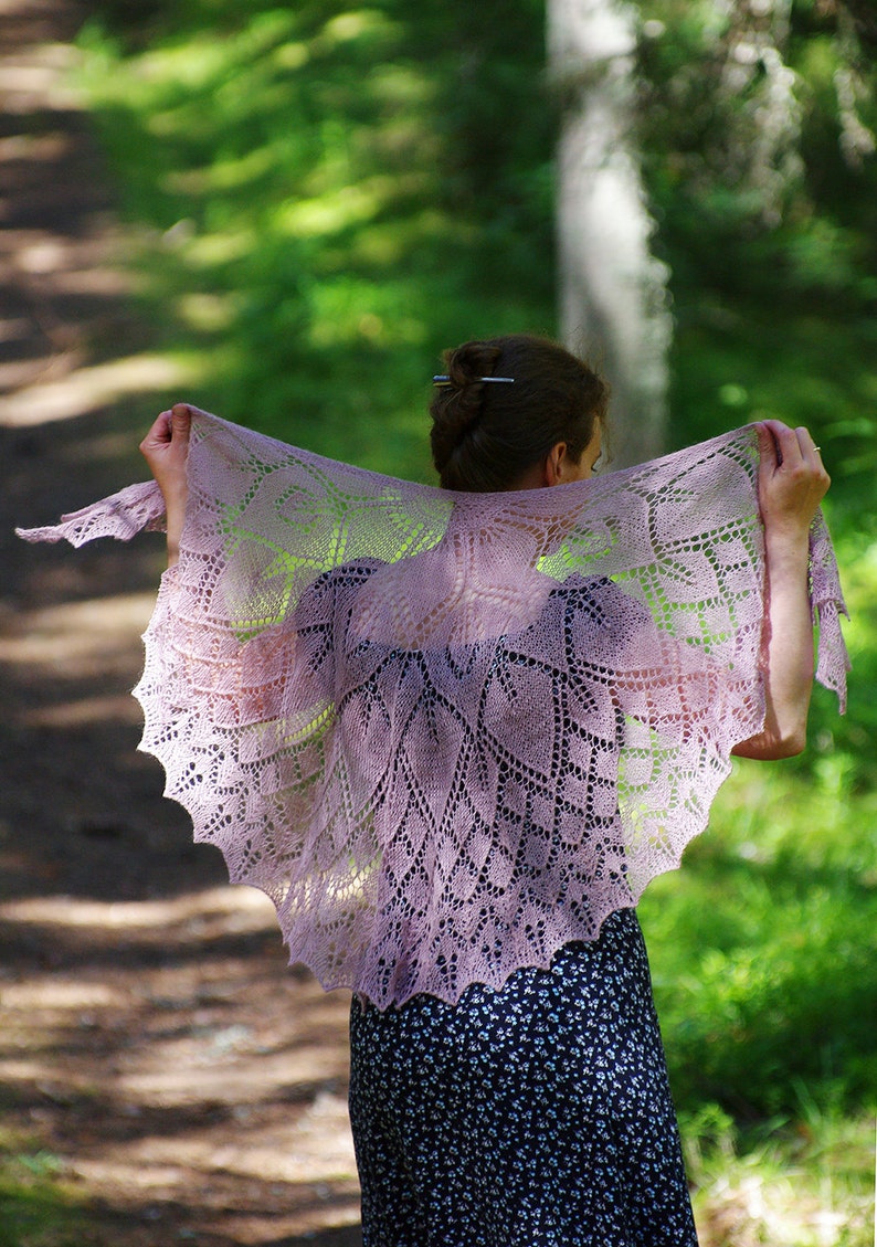 May include: A woman wearing a delicate, lacy, light purple shawl with a floral pattern. The shawl is draped over her shoulders and is flowing in the wind. She is standing in a forest setting.