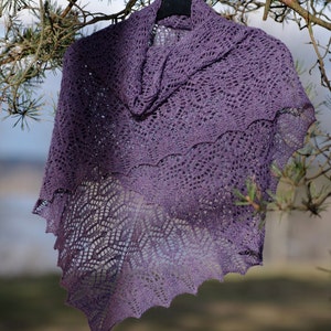 Pode incluir: Um xale de crochê roxo com um padrão de renda está pendurado em um cabide contra um fundo desfocado de galhos de pinheiro e um campo gramado. O texto "PDF Pattern" é visível na parte inferior da imagem.
