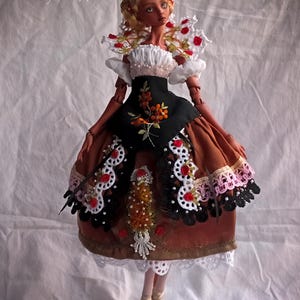 Pode incluir: Uma boneca articulada de porcelana com cabelo loiro, vestindo um corpete preto com bordado floral laranja, uma saia castanha com acabamento em renda branca e preta, detalhes de rosas vermelhas e sapatos dourados. Texto: Amado Gravagno Porcelain Ball jointed dolls.