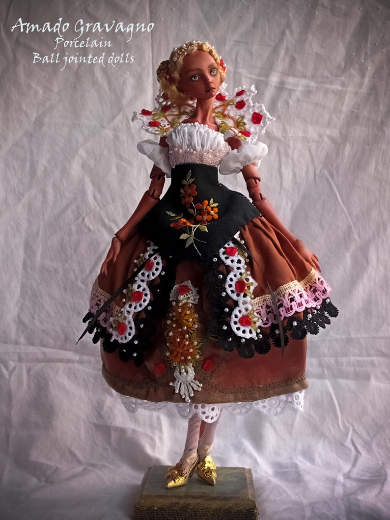 Pode incluir: Uma boneca articulada de porcelana com cabelo loiro, vestindo um corpete preto com bordado floral laranja, uma saia castanha com acabamento em renda branca e preta, detalhes de rosas vermelhas e sapatos dourados. Texto: Amado Gravagno Porcelain Ball jointed dolls.