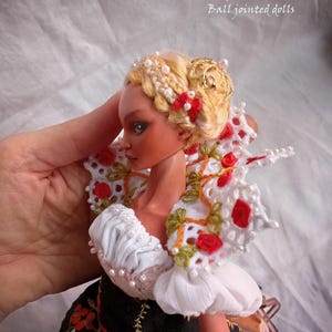 Pode incluir: Uma boneca articulada de porcelana com cabelo loiro preso num penteado com pequenas p&eacute;rolas brancas e um la&ccedil;o vermelho. A boneca usa um top branco com folhos e uma gola de renda com detalhes florais vermelhos e uma saia preta com bordados.