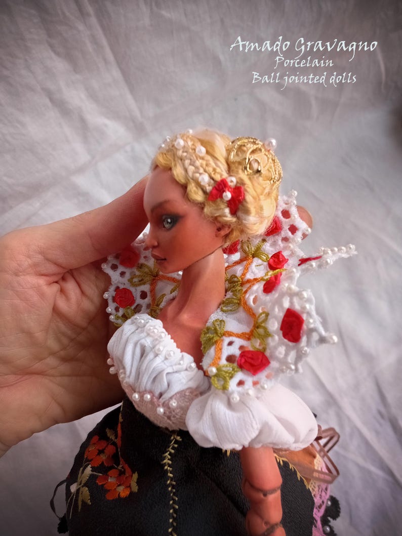 Pode incluir: Uma boneca articulada de porcelana com cabelo loiro preso num penteado com pequenas p&eacute;rolas brancas e um la&ccedil;o vermelho. A boneca usa um top branco com folhos e uma gola de renda com detalhes florais vermelhos e uma saia preta com bordados.
