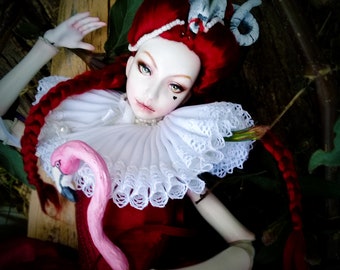 May include: A porcelain ball jointed doll with red hair and a white lace collar. The doll is holding a pink flamingo cane and is wearing a red dress. The doll is lying on a wooden surface with green leaves in the background. The text "Amado Gravagno Porcelain Ball jointed dolls" is visible in the top right corner of the image.