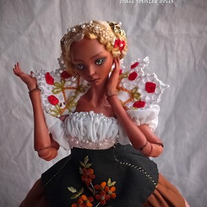 Pode incluir: Uma boneca de porcelana articulada com cabelo loiro tran&ccedil;ado adornado com p&eacute;rolas e la&ccedil;os vermelhos. A boneca usa um top branco com babados e p&eacute;rolas, um grande colarinho de renda branca com detalhes florais vermelhos e um avental preto sobre saia castanha.